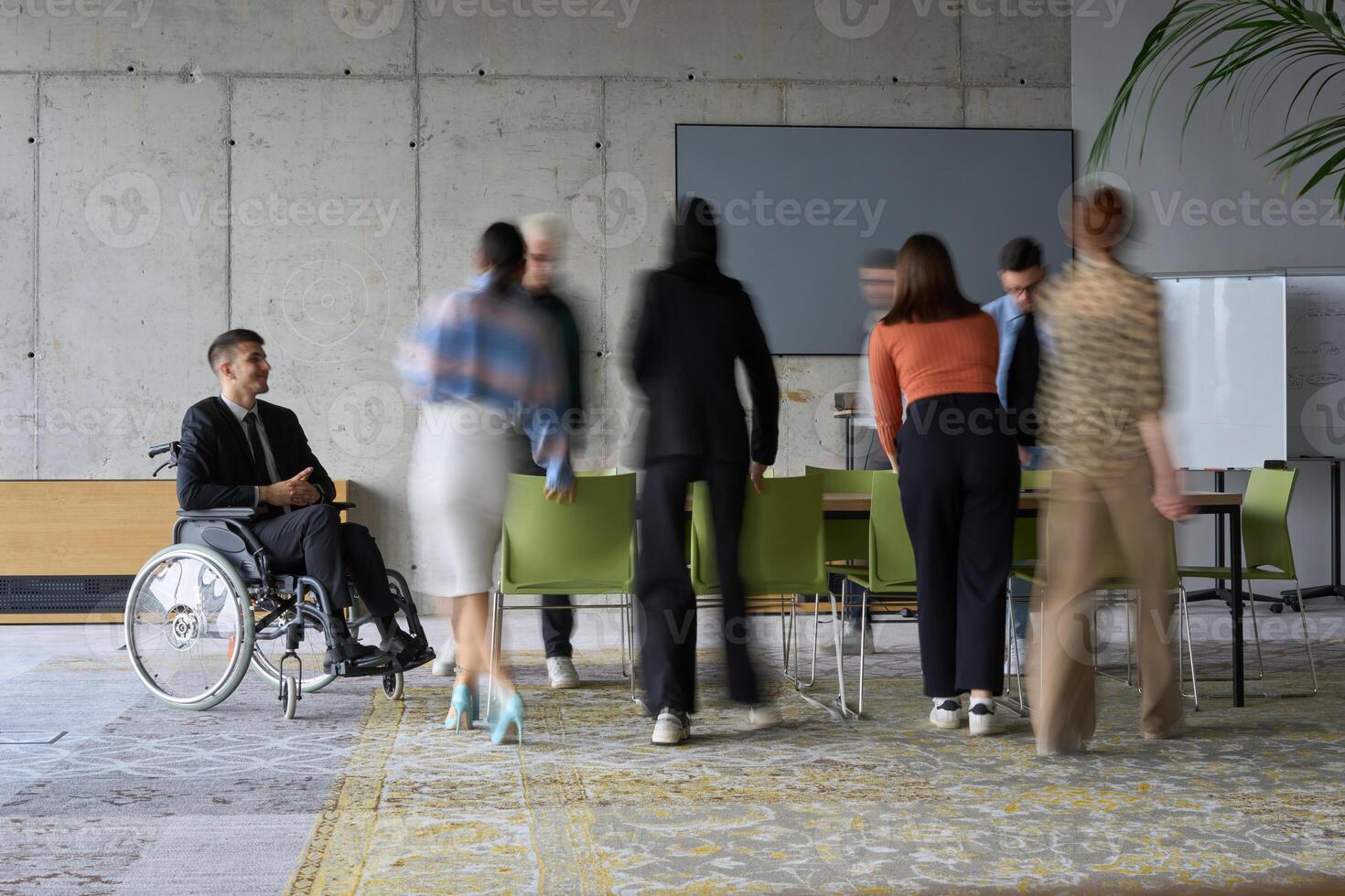 A diverse business team, working urgently around a conference table, collaborates on a meeting while their director, seated in a wheelchair, waits for their input photo
