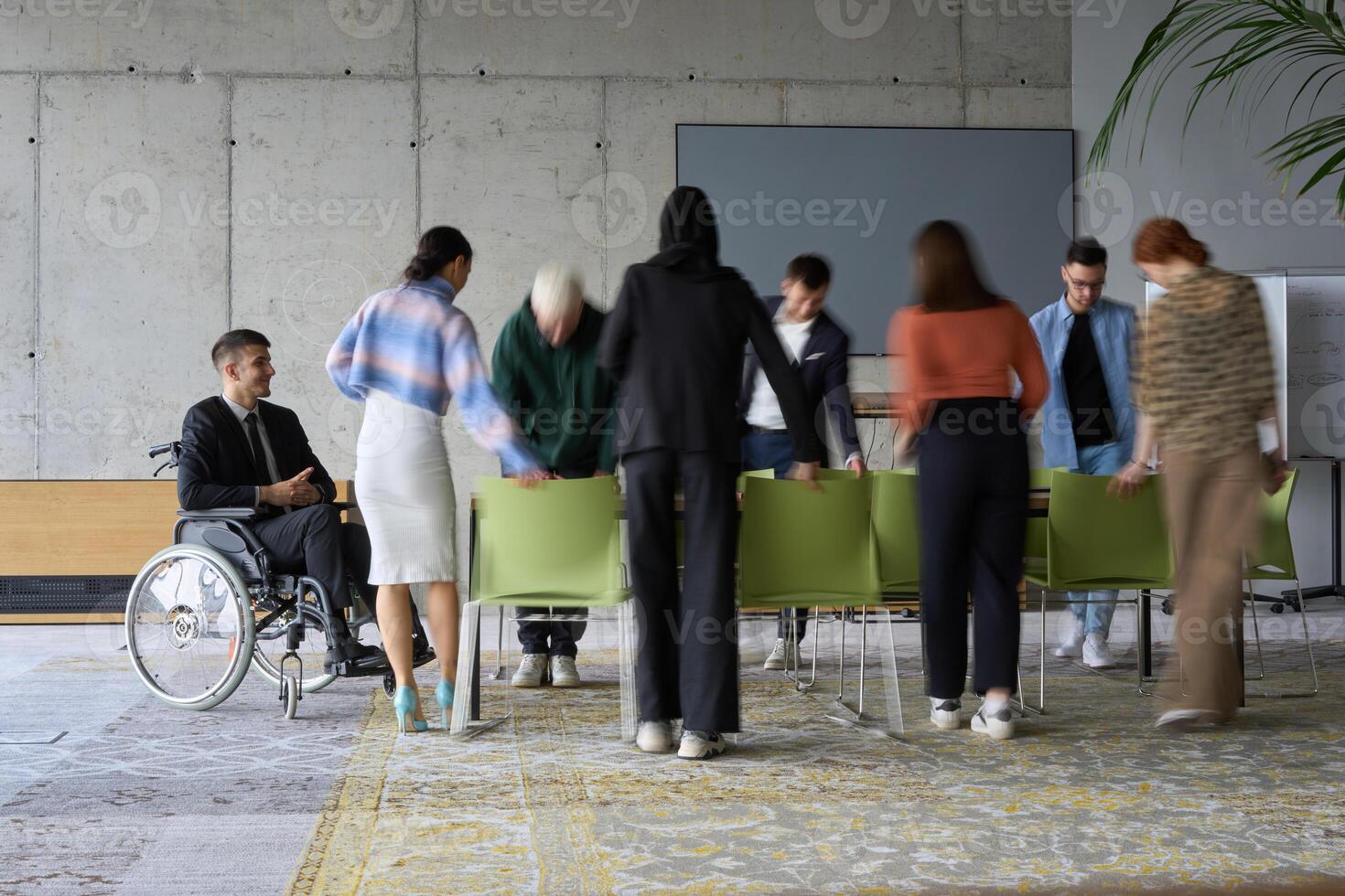 A diverse business team, working urgently around a conference table, collaborates on a meeting while their director, seated in a wheelchair, waits for their input photo