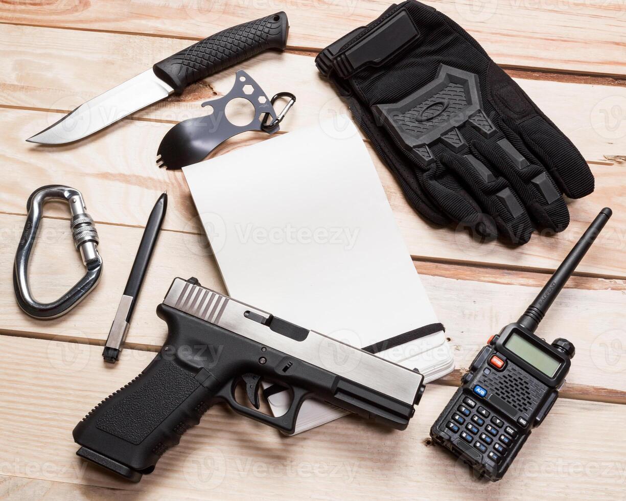 Assault rifle, gun, knife with sheath, compass and notebook with pen on table. photo