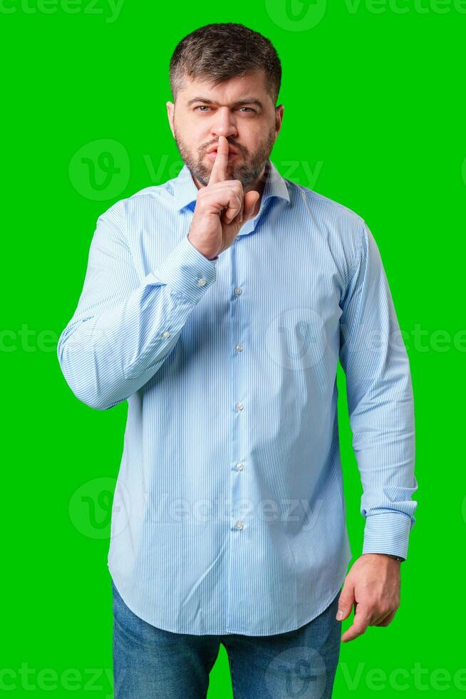A man in a blue shirt gestures for silence with a finger on his lips against a bright green background photo