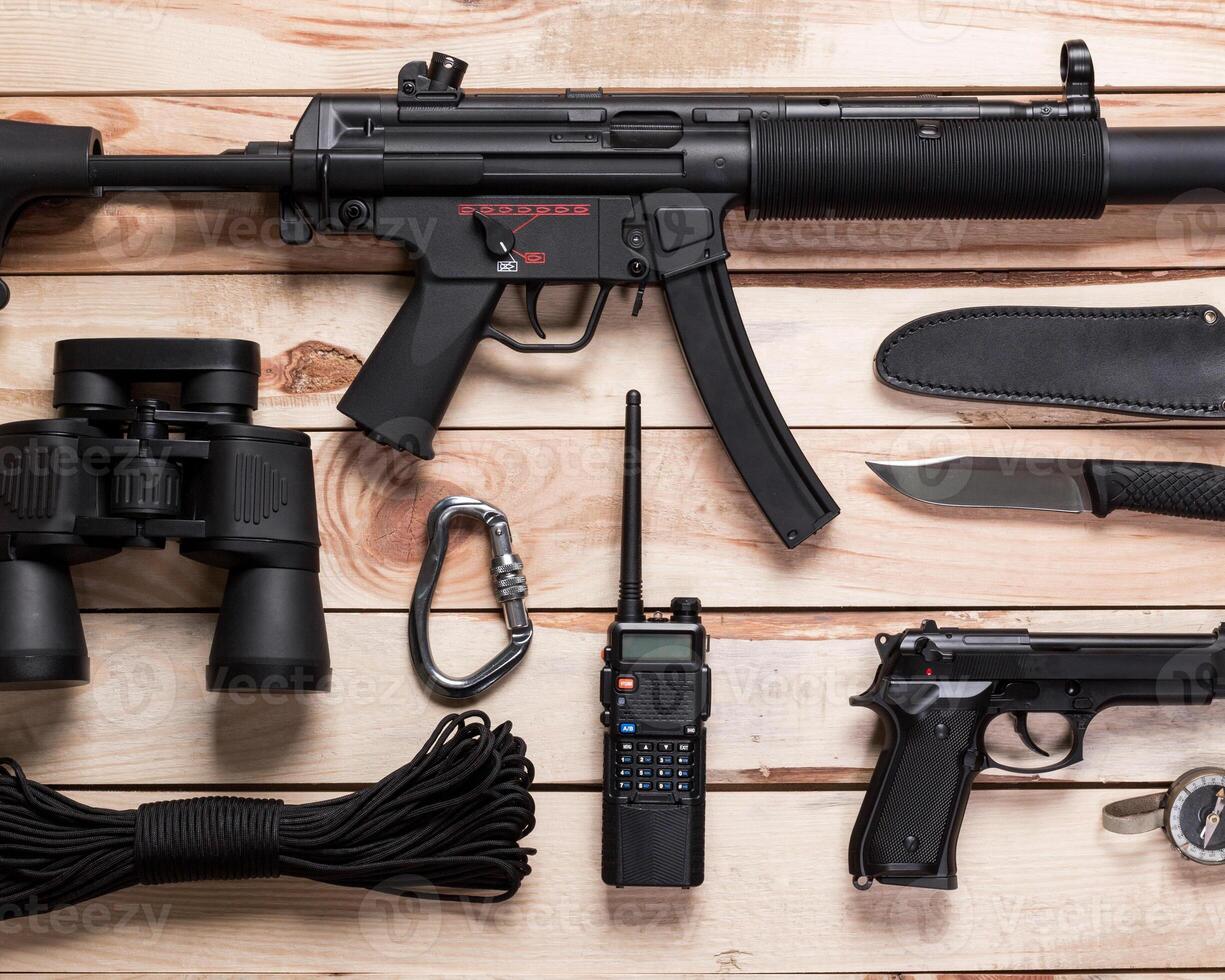 Assault rifle, gun, knife with sheath, compass and notebook with pen on table. photo