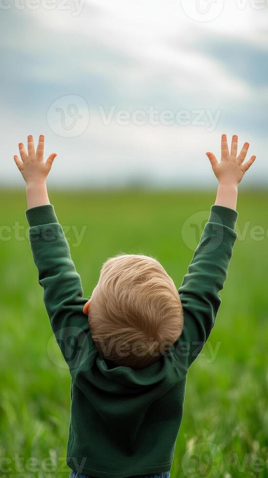A young child joyfully raises hands towards the sky in a lush green field during a bright day photo