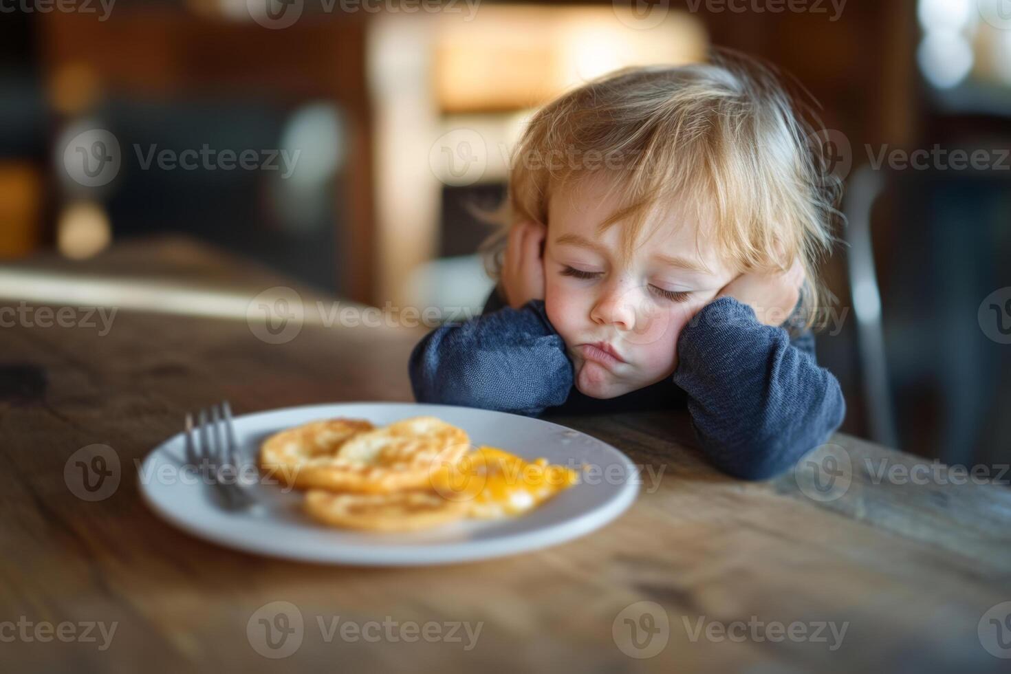 A child looking disappointed while waiting for a meal at a cozy kitchen table in the morning light with a plate of eggs in front photo