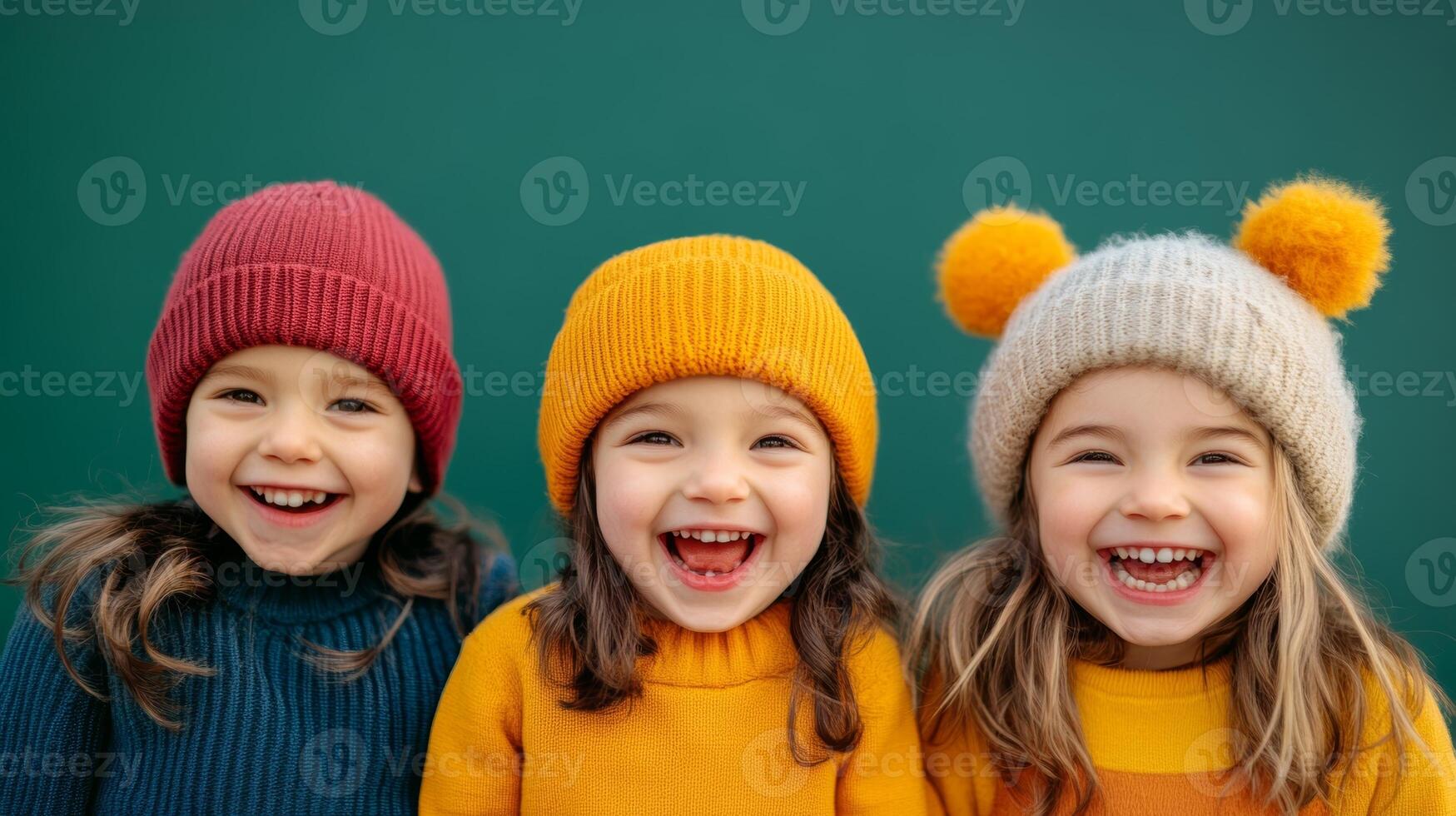 Three young girls wearing colorful knit hats share bright smiles against a teal background during a joyful winter day photo