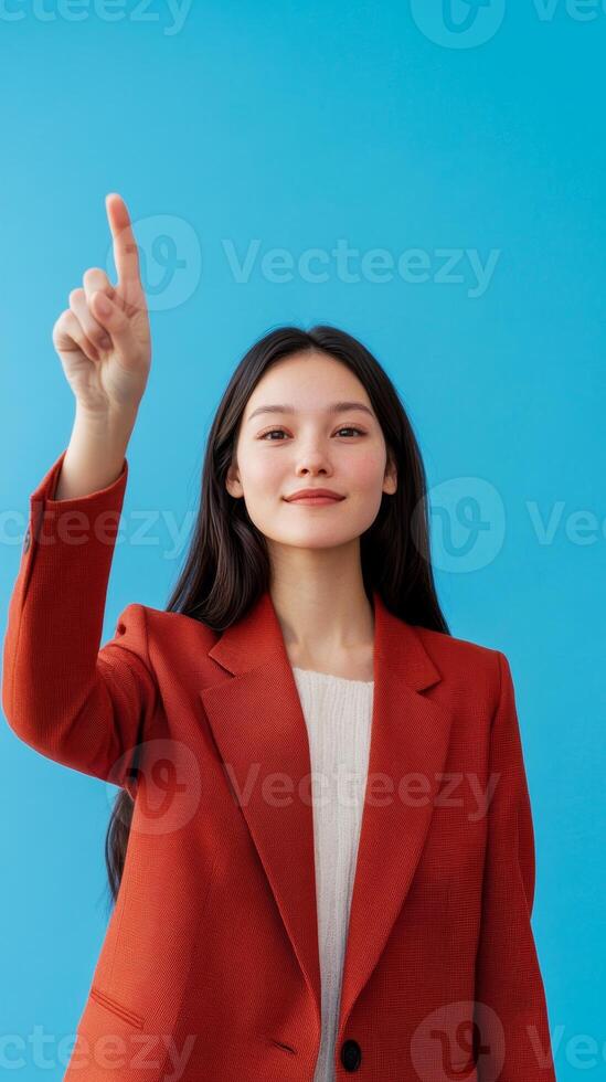 Young woman in a red blazer with long hair pointing up against a vibrant blue background in a bright and cheerful setting photo