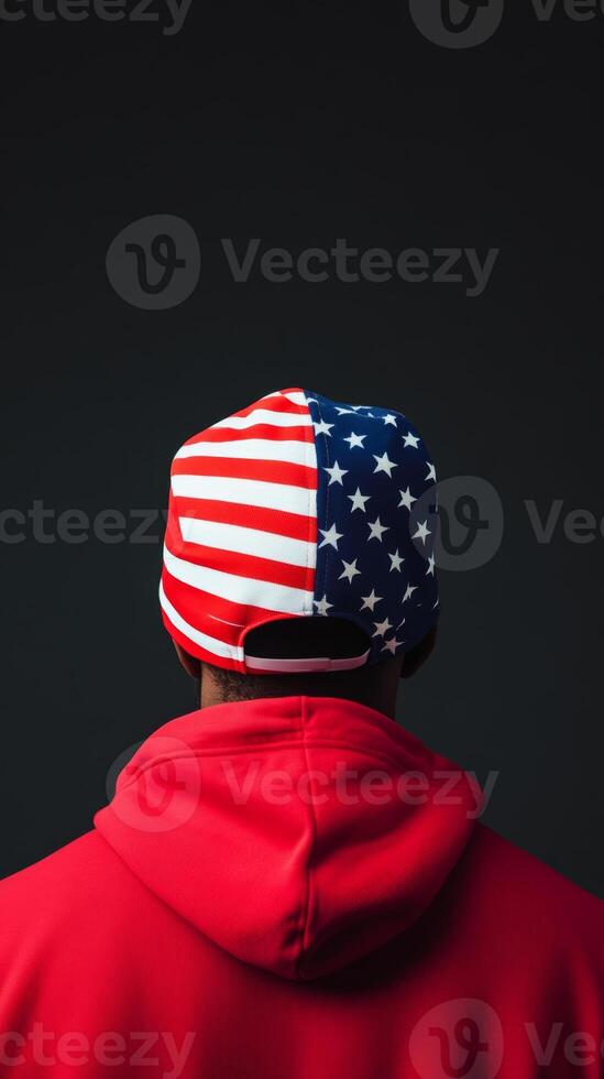 A person wearing a red hoodie and a cap featuring the American flag stands in front of a dark background during a casual indoor setting photo