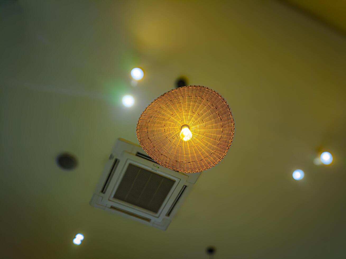 A warm, woven rattan lampshade with a glowing bulb hangs from a ceiling, surrounded by recessed lights and an air conditioning vent, creating a cozy ambiance photo