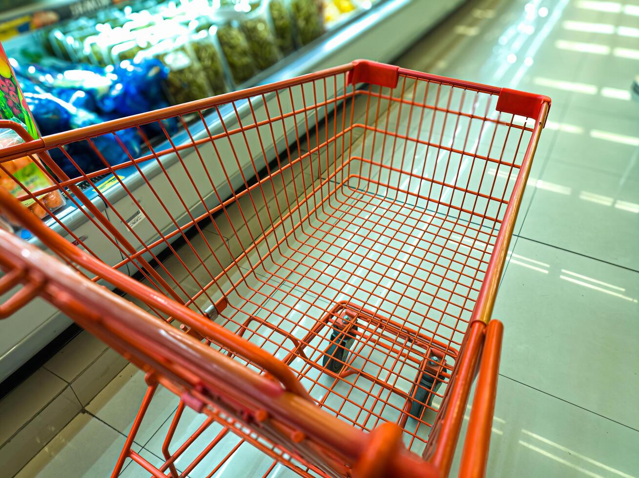 An empty red shopping cart in a grocery store aisle, with shelves of packaged goods in the background. The cart is positioned near the refrigerated section, typical shopping experience photo