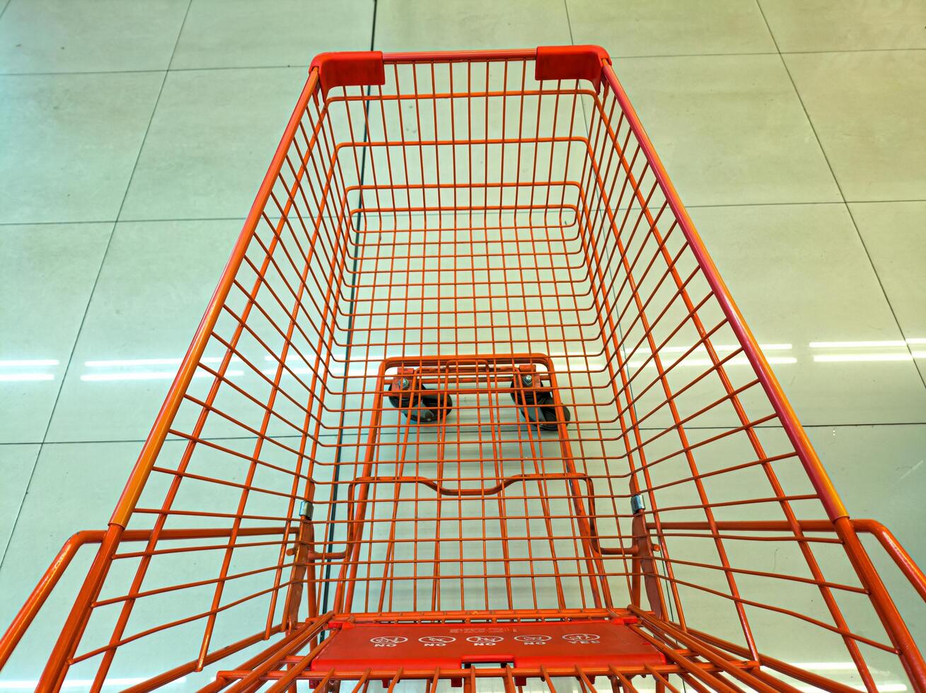 An empty orange shopping cart with a wireframe design is seen from above on a tiled floor. The cart is positioned in a well-lit, clean environment, likely a supermarket or store photo