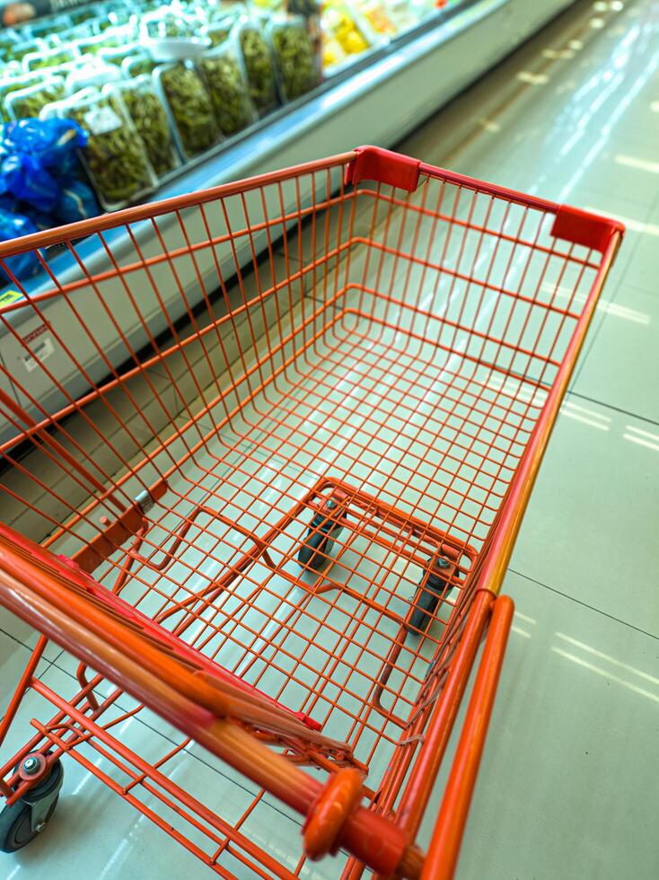 An empty orange shopping cart in a grocery store aisle, with shelves of packaged food items in the background. The cart is positioned near a refrigerated section photo