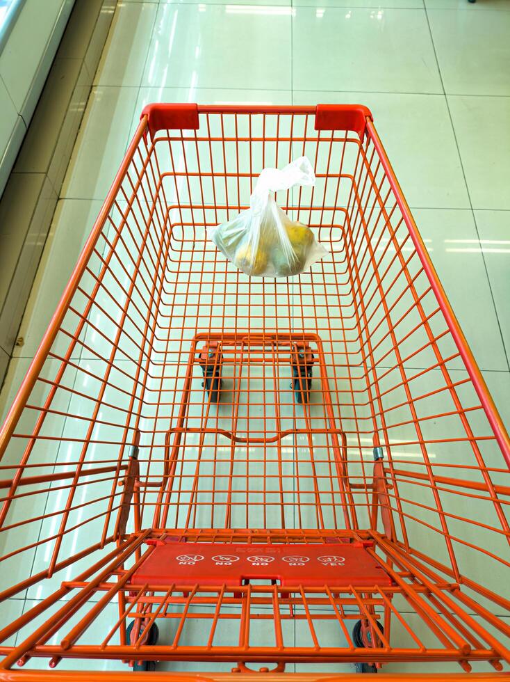 An empty orange shopping cart with a single plastic bag containing fruits, placed in a clean, tiled supermarket aisle photo