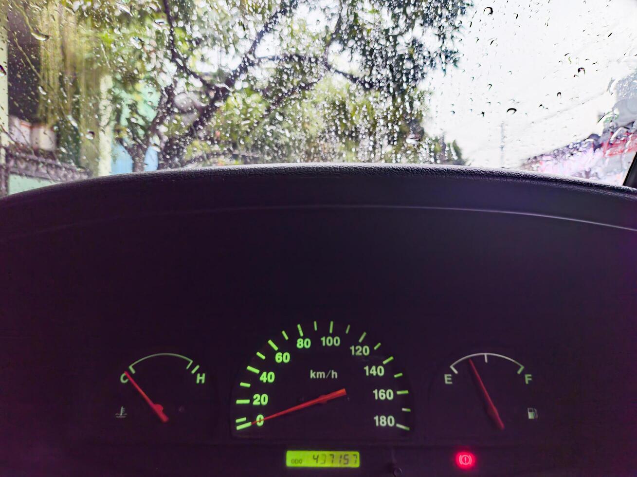 A car dashboard showing speedometer, fuel gauge, and temperature gauge with raindrops on the windshield, indicating a rainy day photo