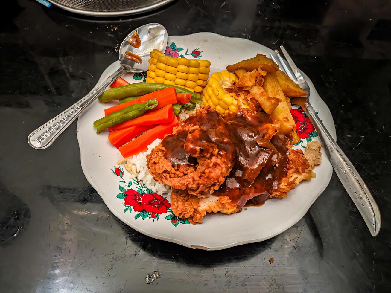 A plate of fried chicken with gravy, rice, corn, green beans, carrots, and fried potatoes on a floral patterned plate, accompanied by a spoon and fork on a black surface photo