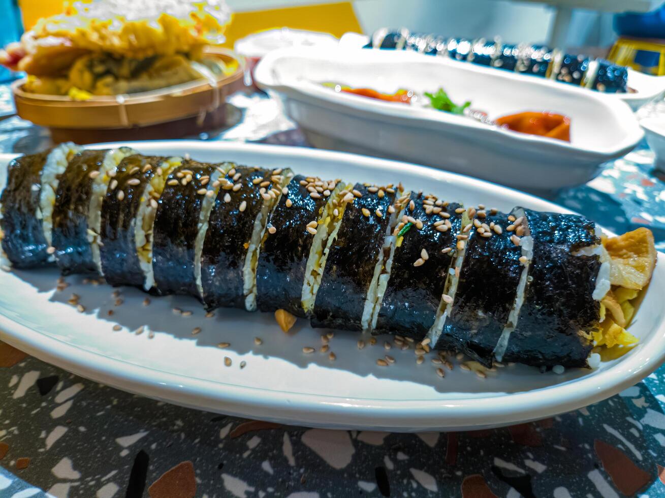 A close up of a sliced Korean gimbap roll on a white plate, garnished with sesame seeds. The background shows other dishes, highlighting a traditional Korean meal setting photo