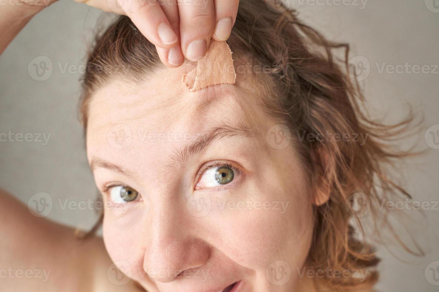 Young caucasian female applying adhesive bandage to forehead wound with smiling expression. photo