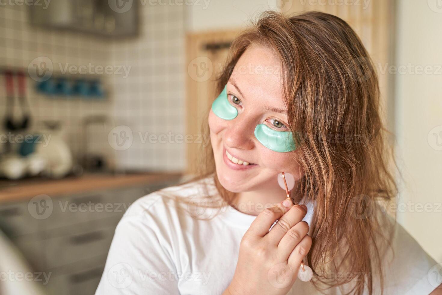Young caucasian female with eye patches using jade roller in cozy kitchen setting for skincare routine. photo