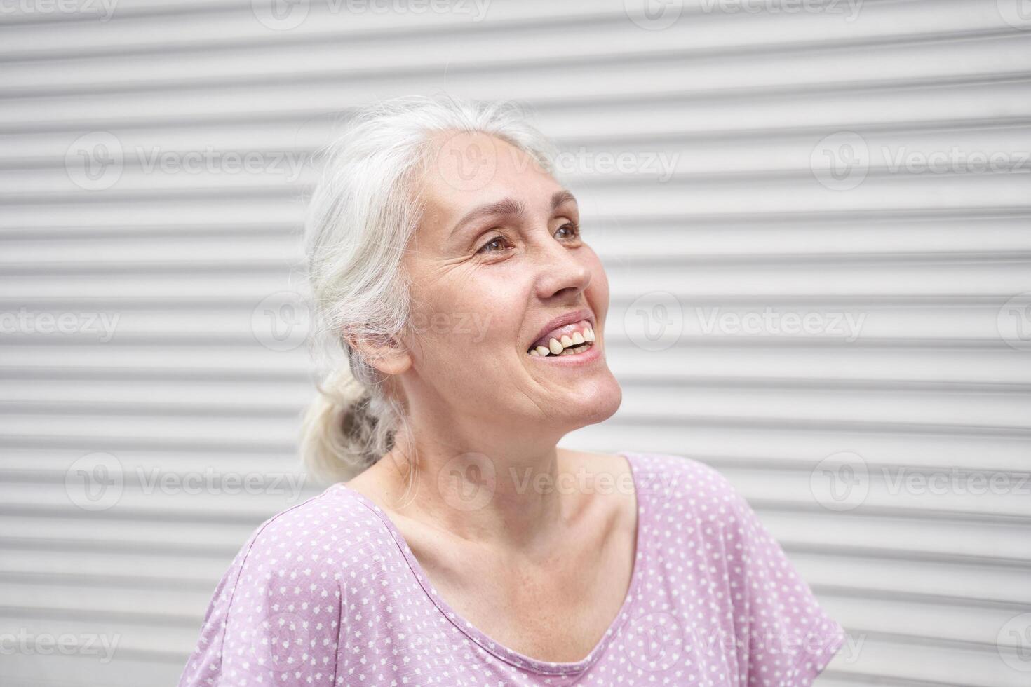 Elderly caucasian woman smiling joyfully in light pink polka dot top against white striped background. photo
