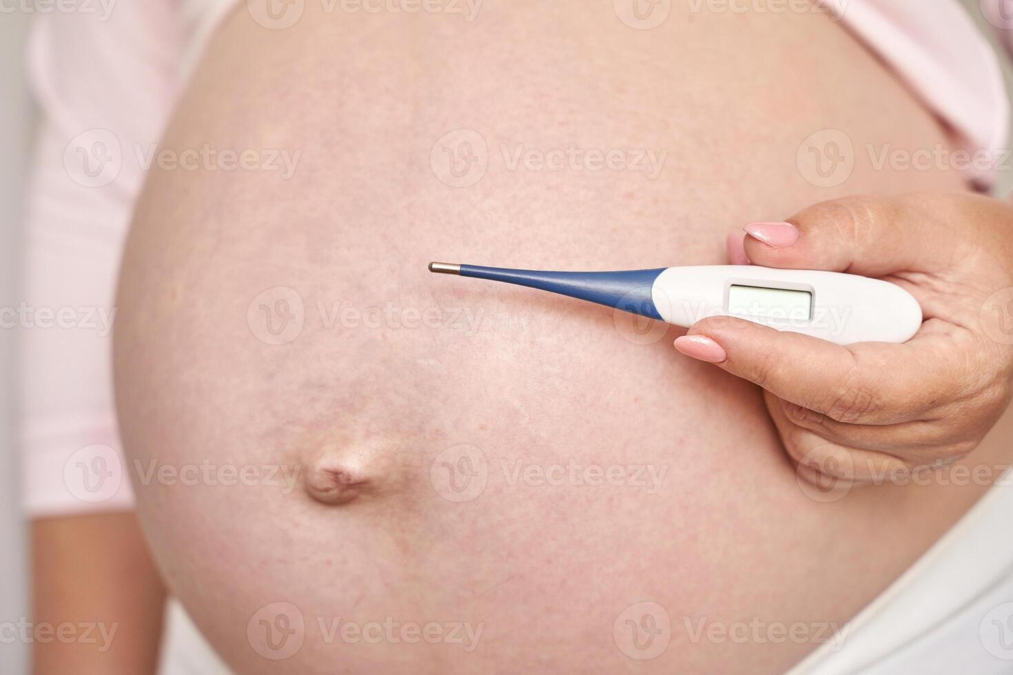 Pregnant caucasian woman holding digital thermometer on bare belly in close up maternity health image. photo