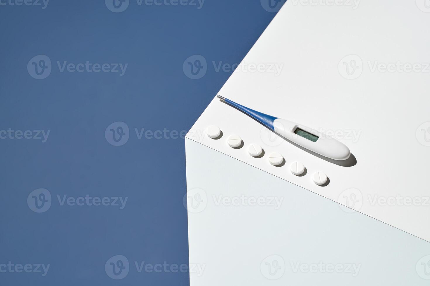Digital thermometer and pills on minimalist white surface against blue background. photo