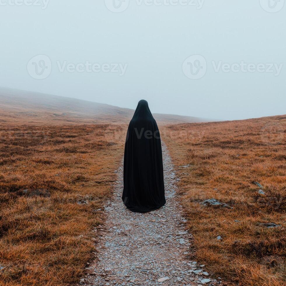 Mysterious figure in black robe walks down a path in a foggy landscape photo