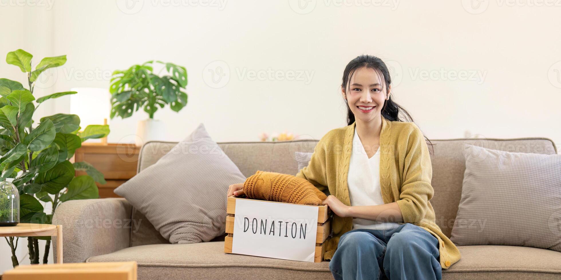 Smiling woman sitting on the couch with a donation box, proudly presenting folded clothes in a cozy living room photo