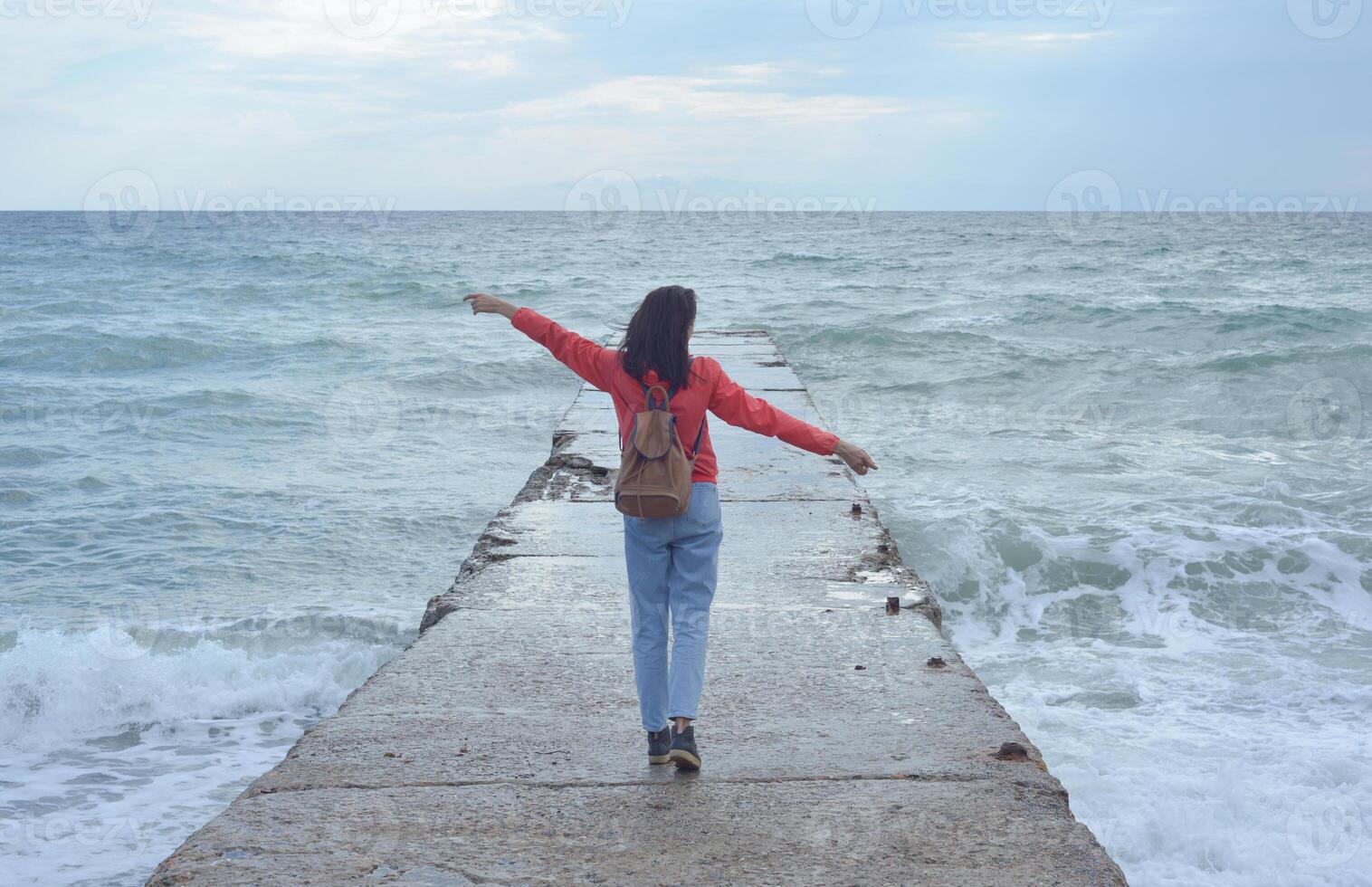 Rear view of a young woman tourist with a backpack standing on the pier and waves hitting the pier in a storm. City of Sudak, Crimea. photo