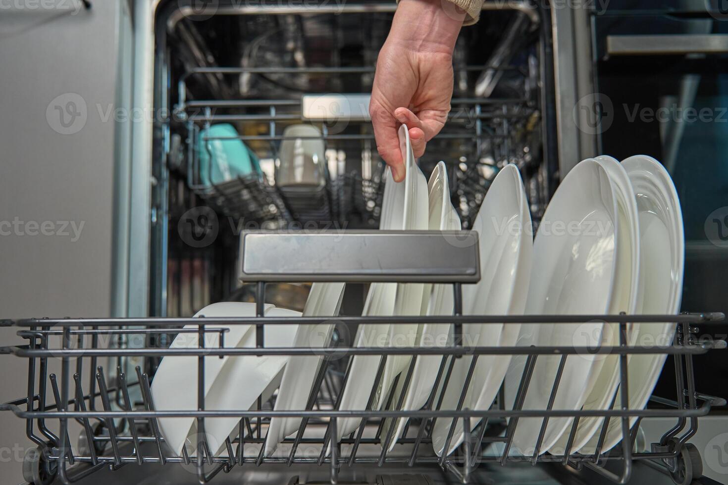 Woman unloading clean white plates from the bottom rack of dishwasher in kitchen. Person performing everyday household chores, using modern home appliance photo