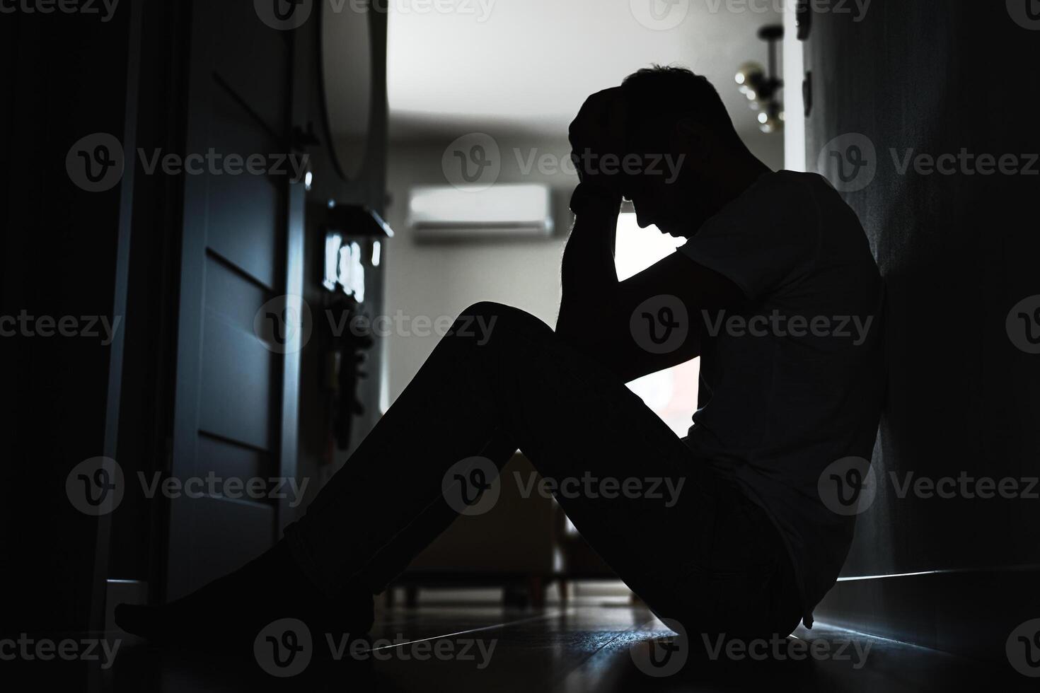 Depressed young man sitting on floor in dark room. Silhouette of young male suffering from loneliness and anxiety, covering his face with hands. Person with mental disorder photo