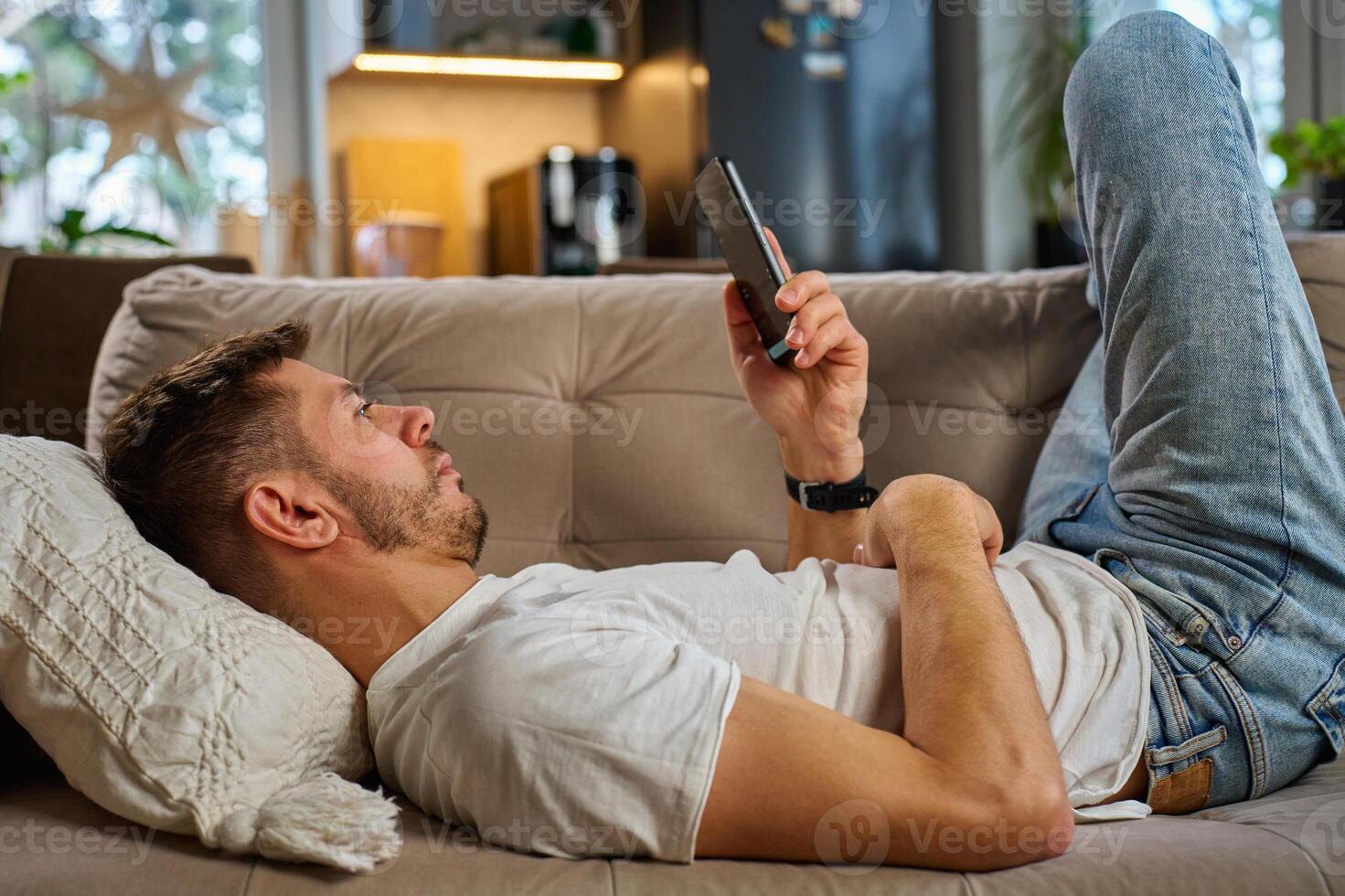 Man in white t-shirt and jeans relaxing on beige sofa, using his smartphone in living room. Male resting on couch browsing social media on mobile phone photo