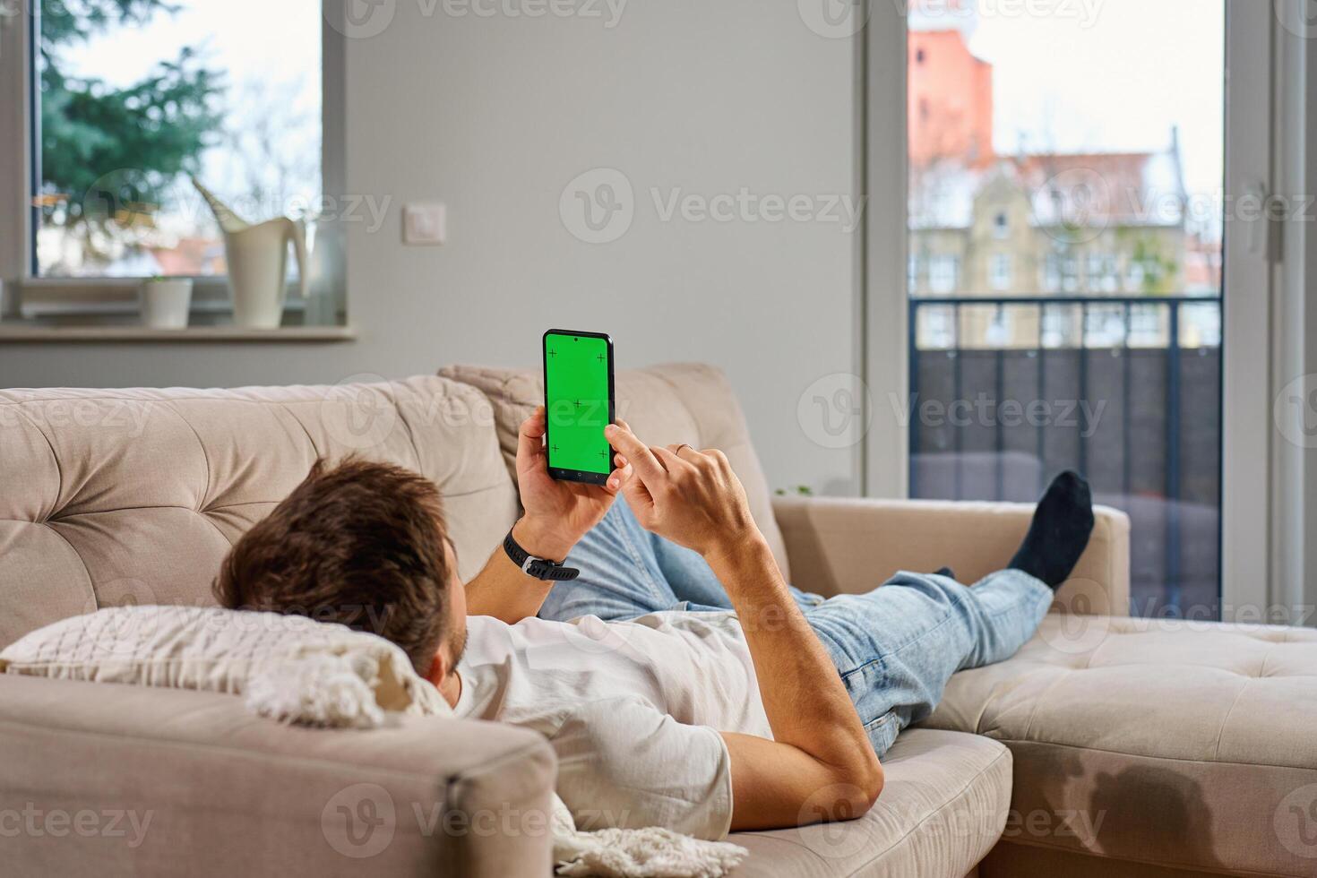 Man in casual clothes lies on beige couch, holding smartphone with green screen, relaxing in modern living room. Male resting on sofa and watching online on mobile phone with chroma key display photo