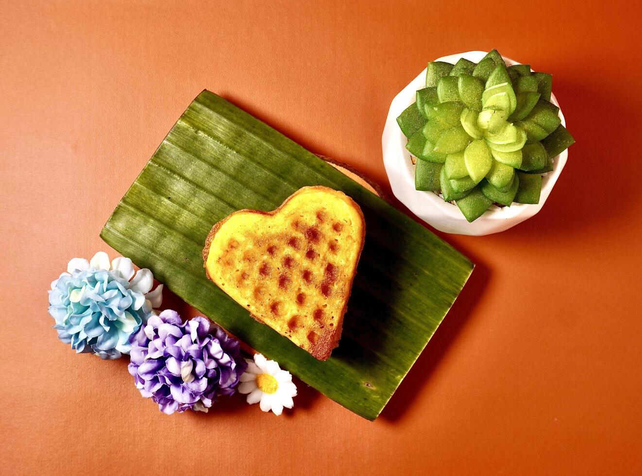 Heart shaped waffle cake. Kue Bapel hati. Sweet dessert snack isolated on horizontal orange brown terracota background from top angle view with potted cactus and flowers plant decoration. photo