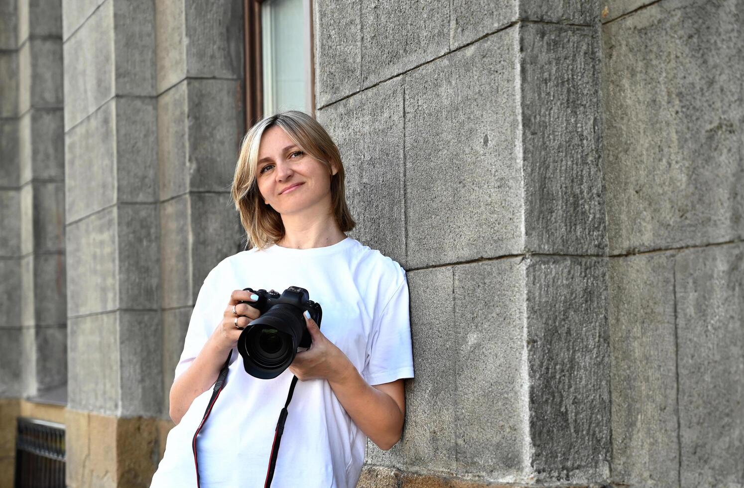 This is a portrait of a woman holding a DSLR camera with a stone wall of a building as a backdrop. She is looking to her left and has a soft smile. photo