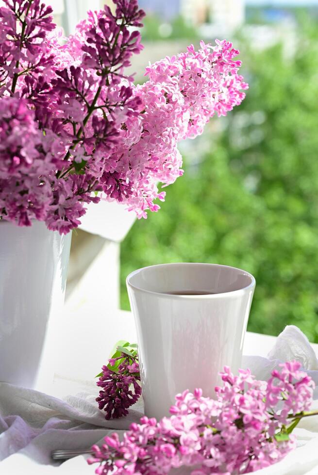 The postcard is beautifulon a shunny day. A mug of tea, an old book and a bouquet of purple lilac. A beautiful still life. Spring time. The concept of Good morning. photo