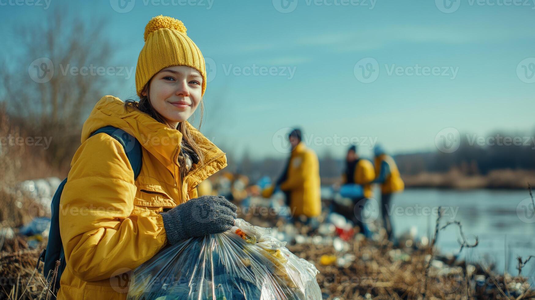 A group of volunteers gathers along a riverbank, collecting trash under a clear blue sky. One young girl smiles as she holds a bag filled with litter. The atmosphere is positive and community-driven photo
