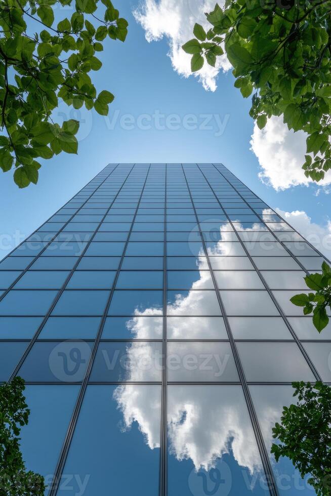 Looking up at a sleek glass building, vibrant green leaves frame the view, while fluffy white clouds reflect off the shiny surface beneath a clear blue sky. photo