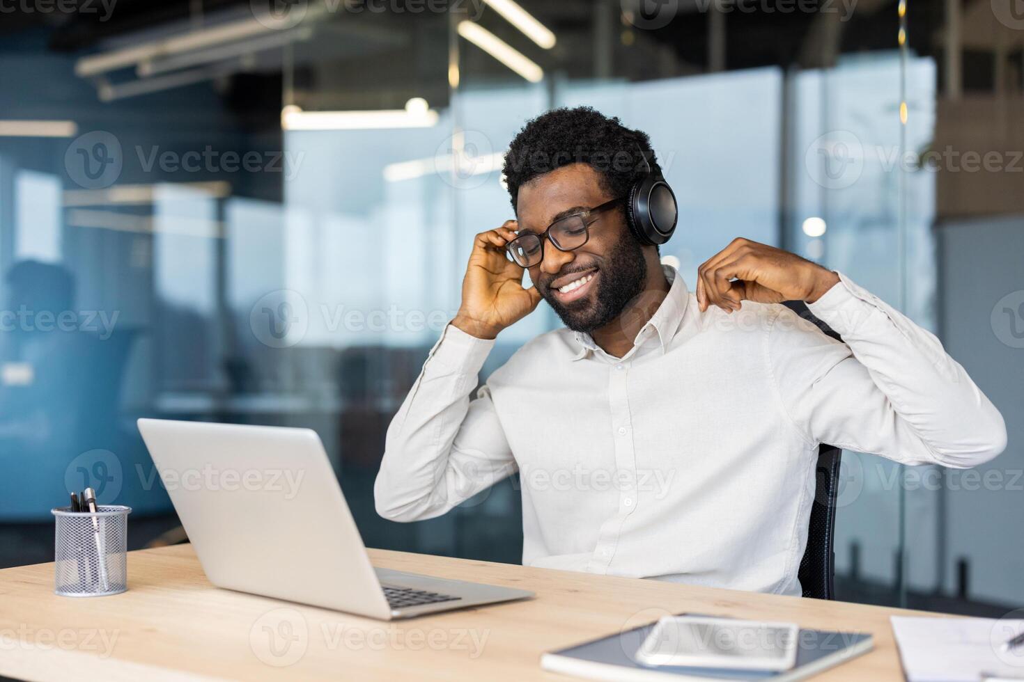 Young man at workplace working with laptop, listening to music, smiling and singing along. Worker in headphones happily resting and relaxing while working in the evening. photo