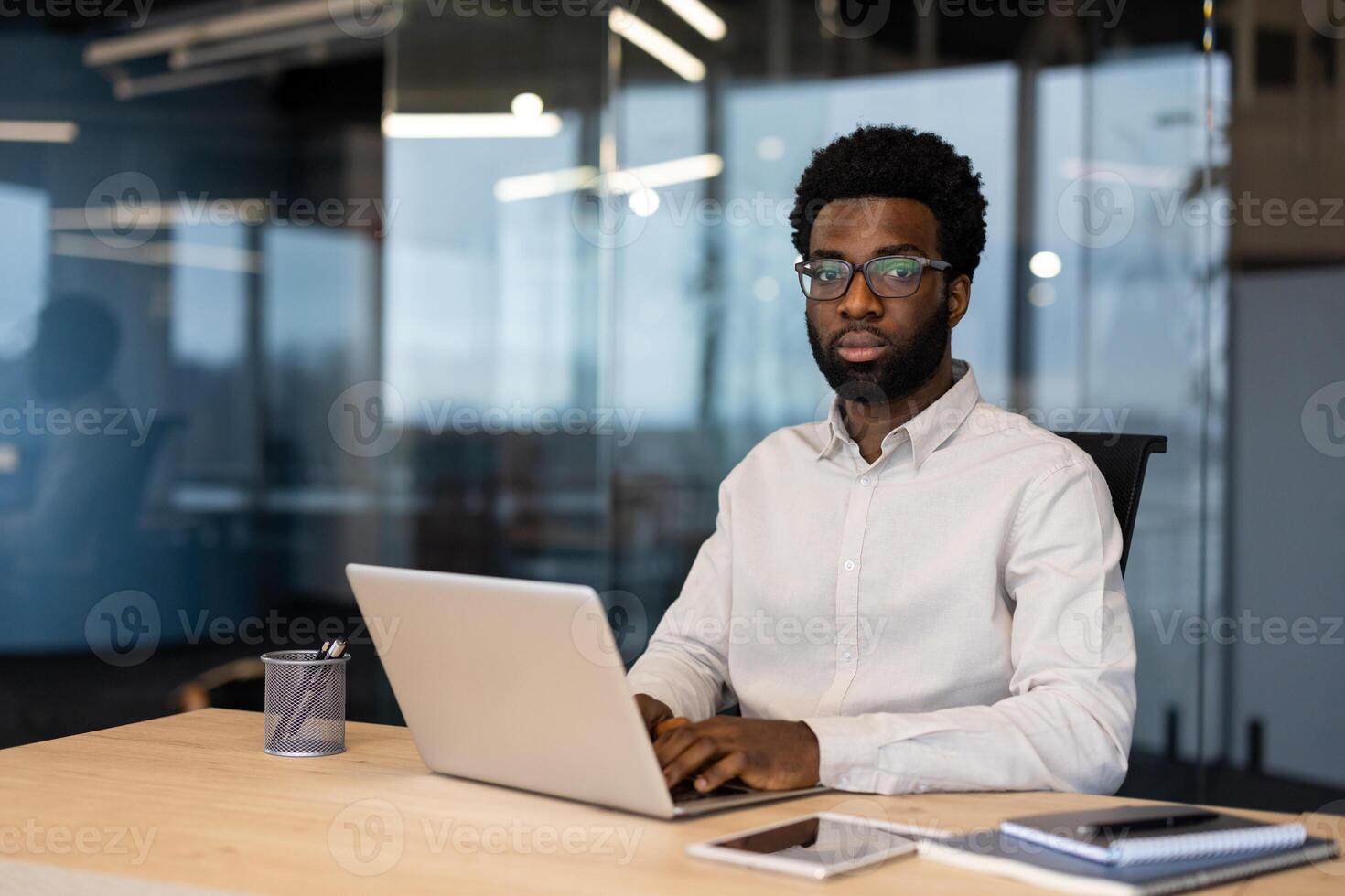 Portrait of a young serious man inside the office with a laptop. Businessman looking at the camera, working in the evening at the workplace. photo