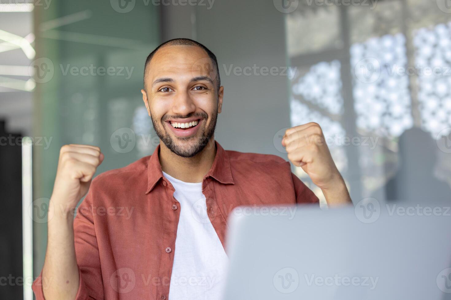 A cheerful individual in an office environment expressing joy with raised fists, representing success and positivity. The light-filled background complements sense of accomplishment conveyed. photo
