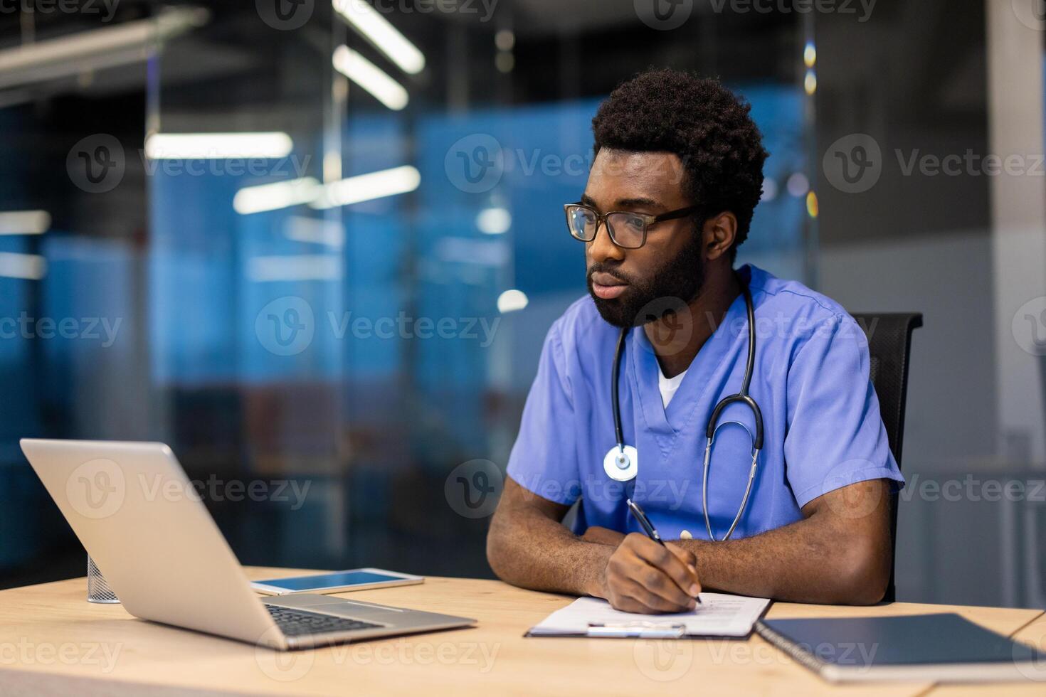 Serious thinking doctor working inside clinic office, man in medical gown with stethoscope writing data in patient treatment report, man working in evening with laptop. photo