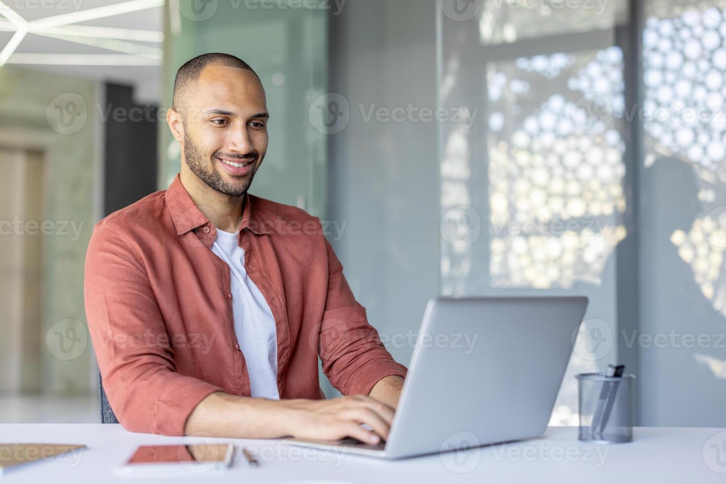 A professional man using a laptop while seated at a desk in a bright, modern workspace, exuding positivity and focus. The scene highlights productivity, business, and a pleasant working atmosphere. photo