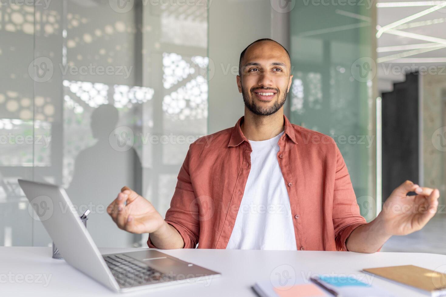 A young man in a casual shirt smiling and meditating at a desk with a laptop in a bright and modern office environment. The mood conveys calmness and balance during work hours. photo