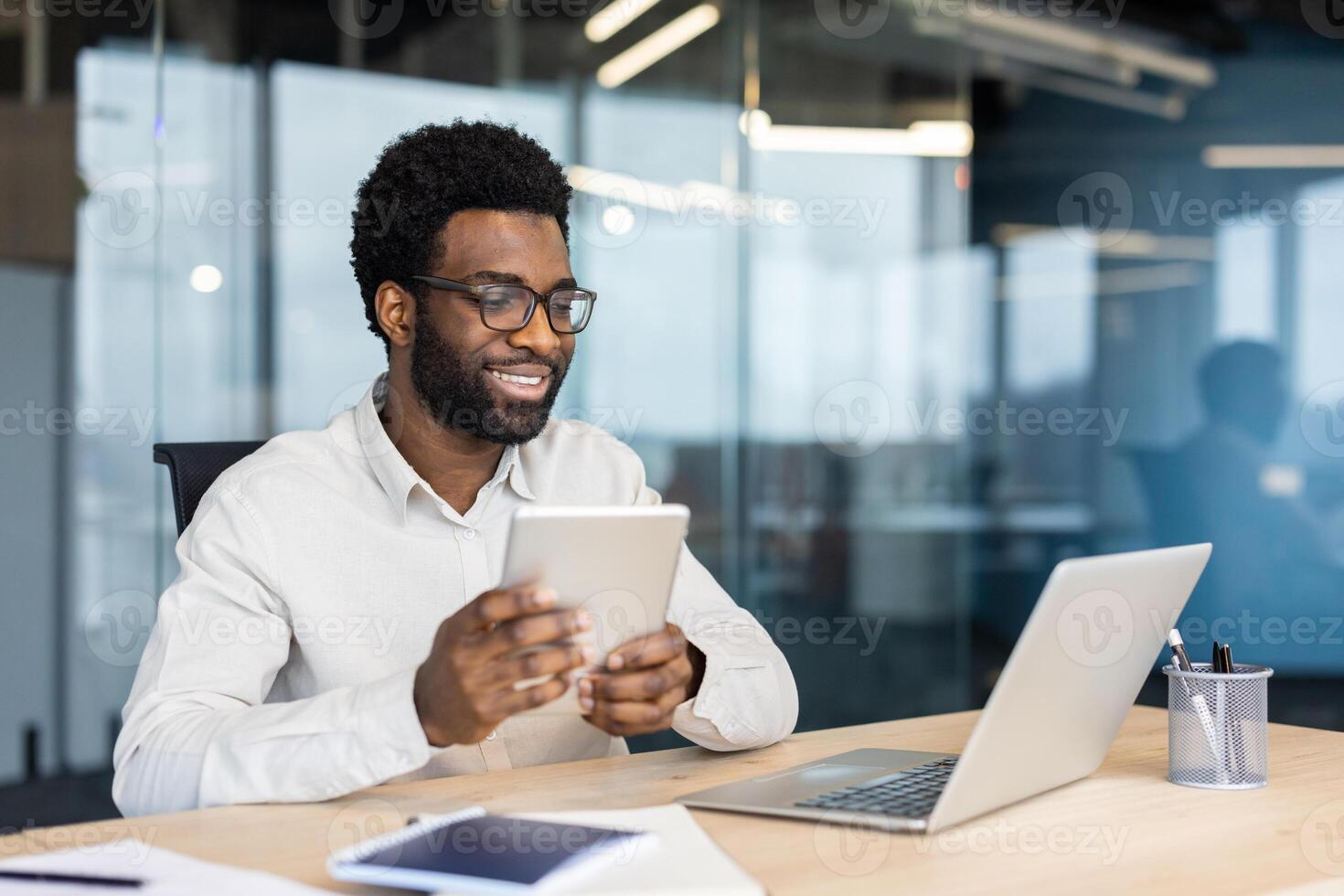 Successful businessman smiling working with laptop inside office at workplace, man holding tablet computer with satisfaction in hands, young developer, financier investor. photo