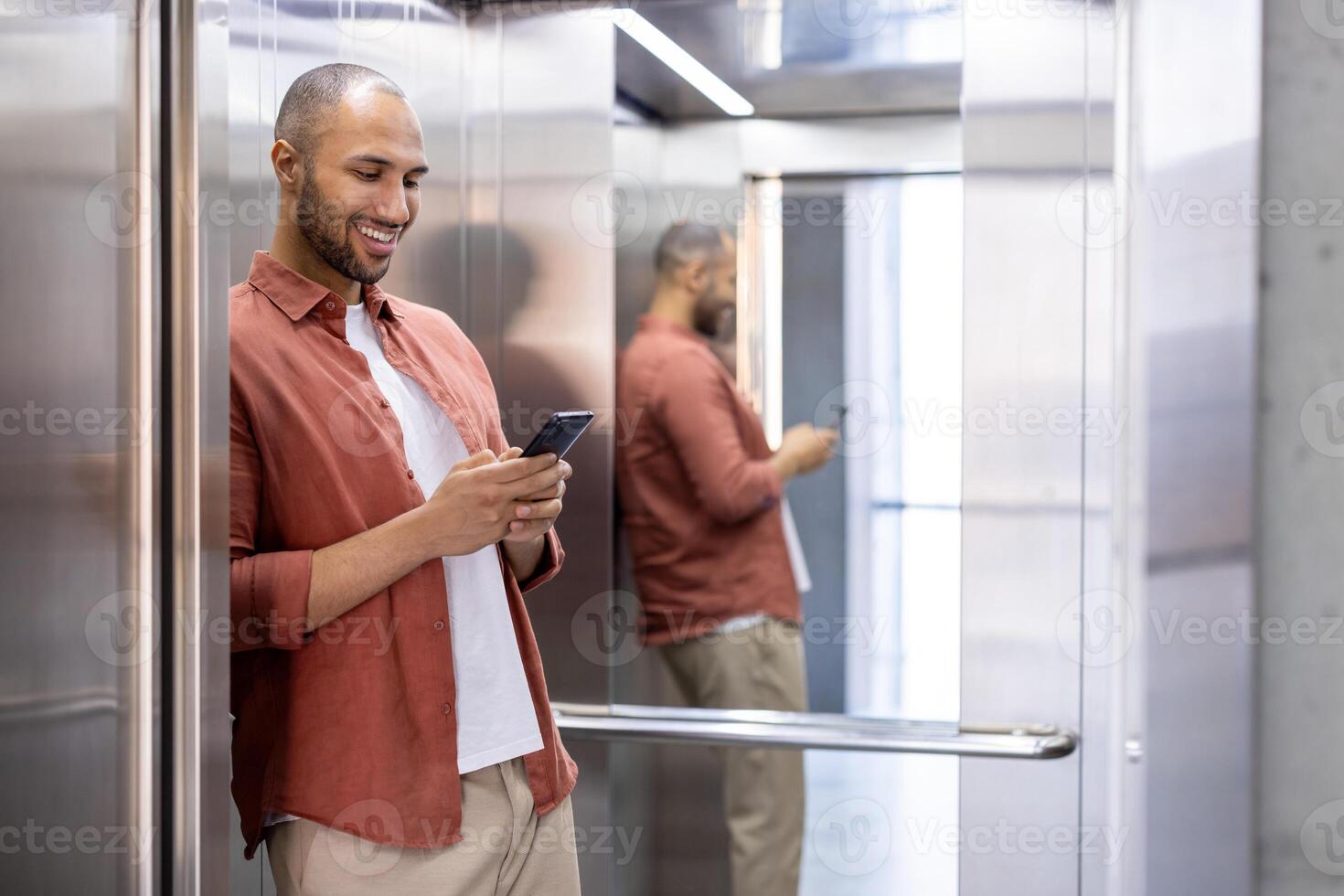 Man standing in elevator using smartphone, leaning against mirror wall, smiling effortlessly, dressed in casual attire. Scene depicts connectivity, communication, modern technology, daily routine. photo