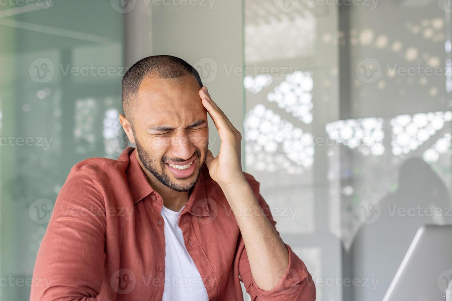 A man sitting indoors with a pained expression, holding his head as if experiencing a headache or stress, dressed casually and with a modern context suggesting work or personal challenge. photo