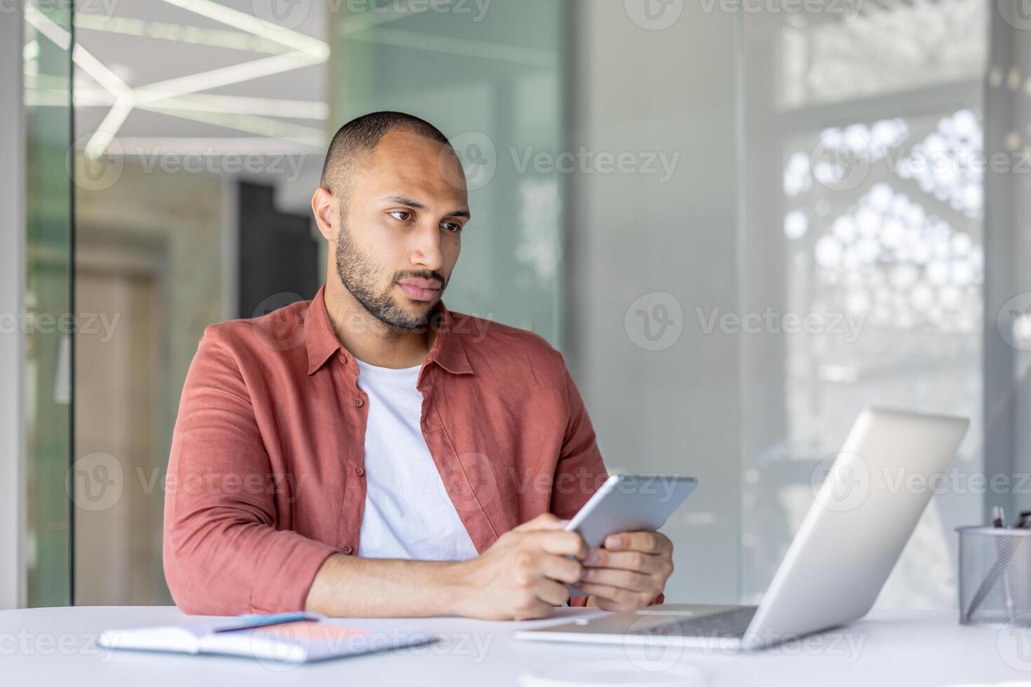 A man holds a tablet while working at a desk with a laptop and notebook in a contemporary office space. He appears focused and immersed in his task. photo