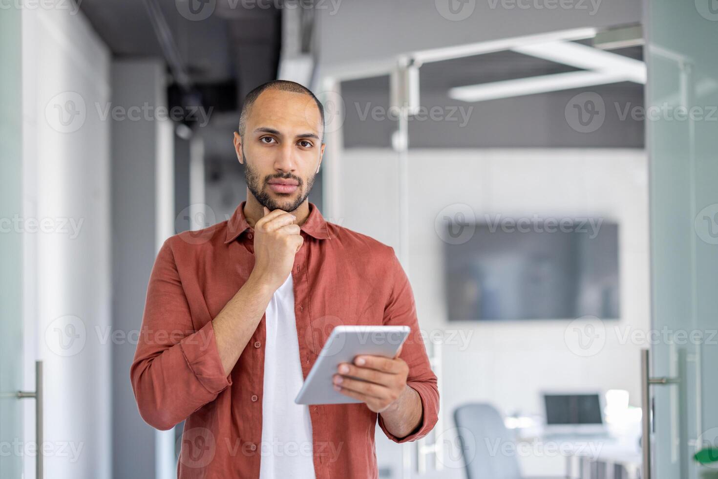 Portrait of a serious confident man with a tablet computer in his hands, businessman looking at the camera, standing inside the office at his workplace in casual clothes. photo