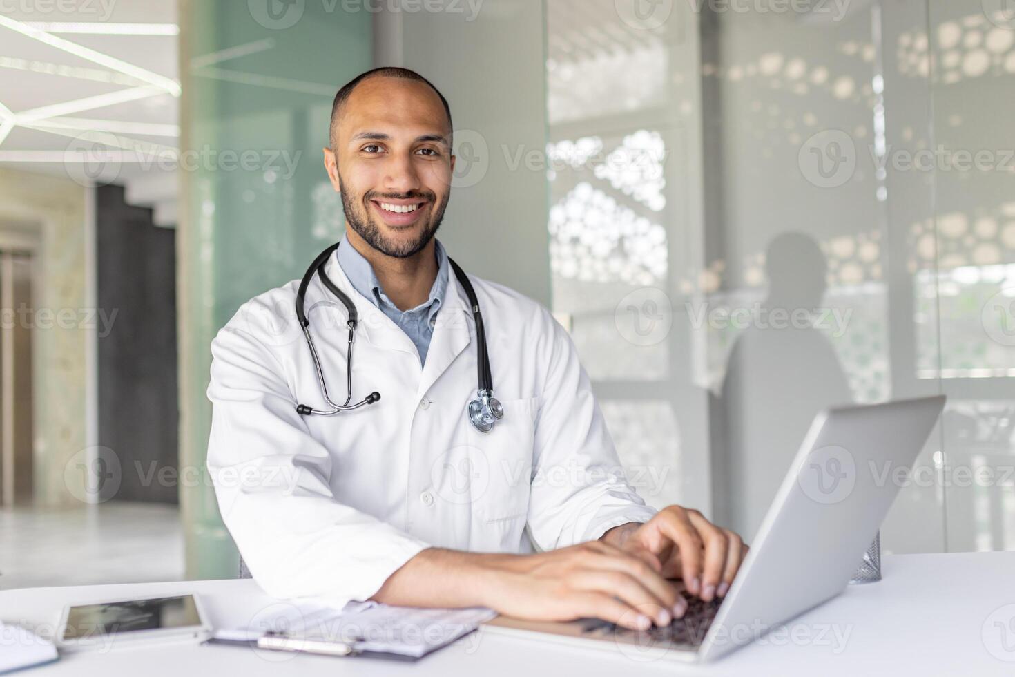 Portrait of a successful doctor in a white medical coat, the man is smiling and looking at the camera, the man is working inside the office with a laptop, consulting patients remotely. photo