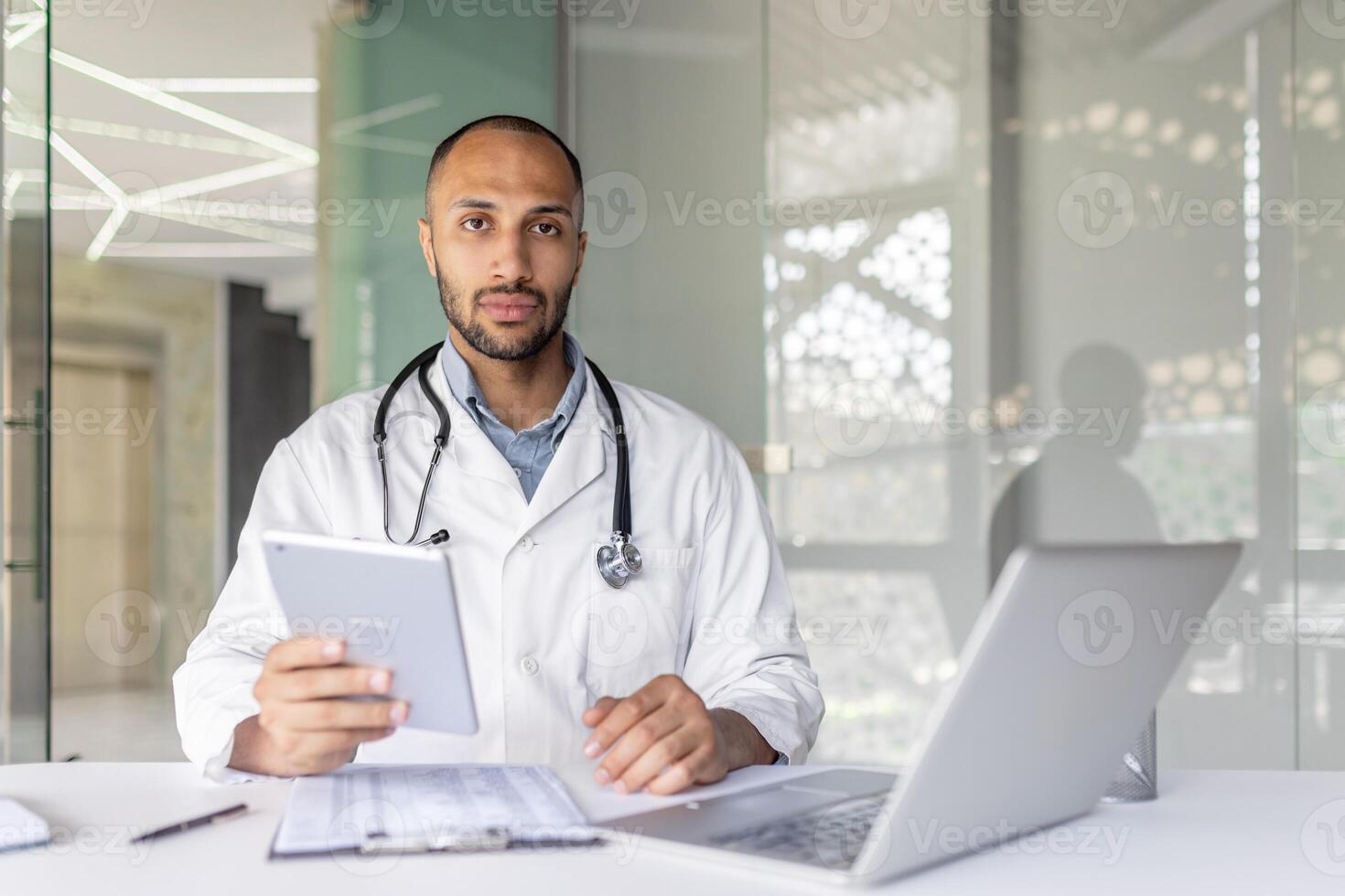 Medical professional dressed in a white coat working with a tablet and computer. The setting highlights professionalism and modern healthcare practices. The focus is on technology and consultations. photo