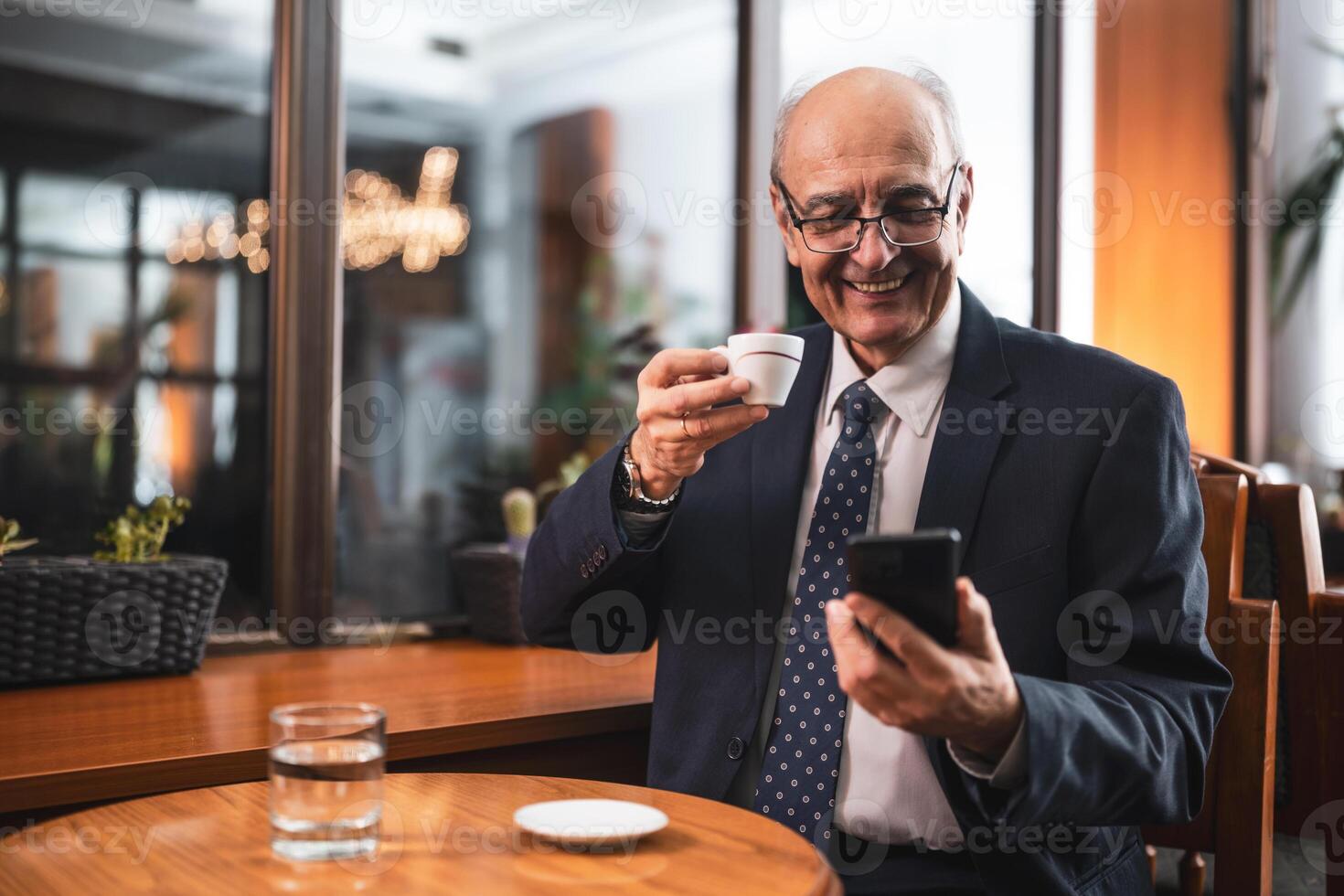 Cheerful senior businessman with eyeglasses in elegant suit using mobile phone and drinking coffee while sitting in the cozy city cafe. photo