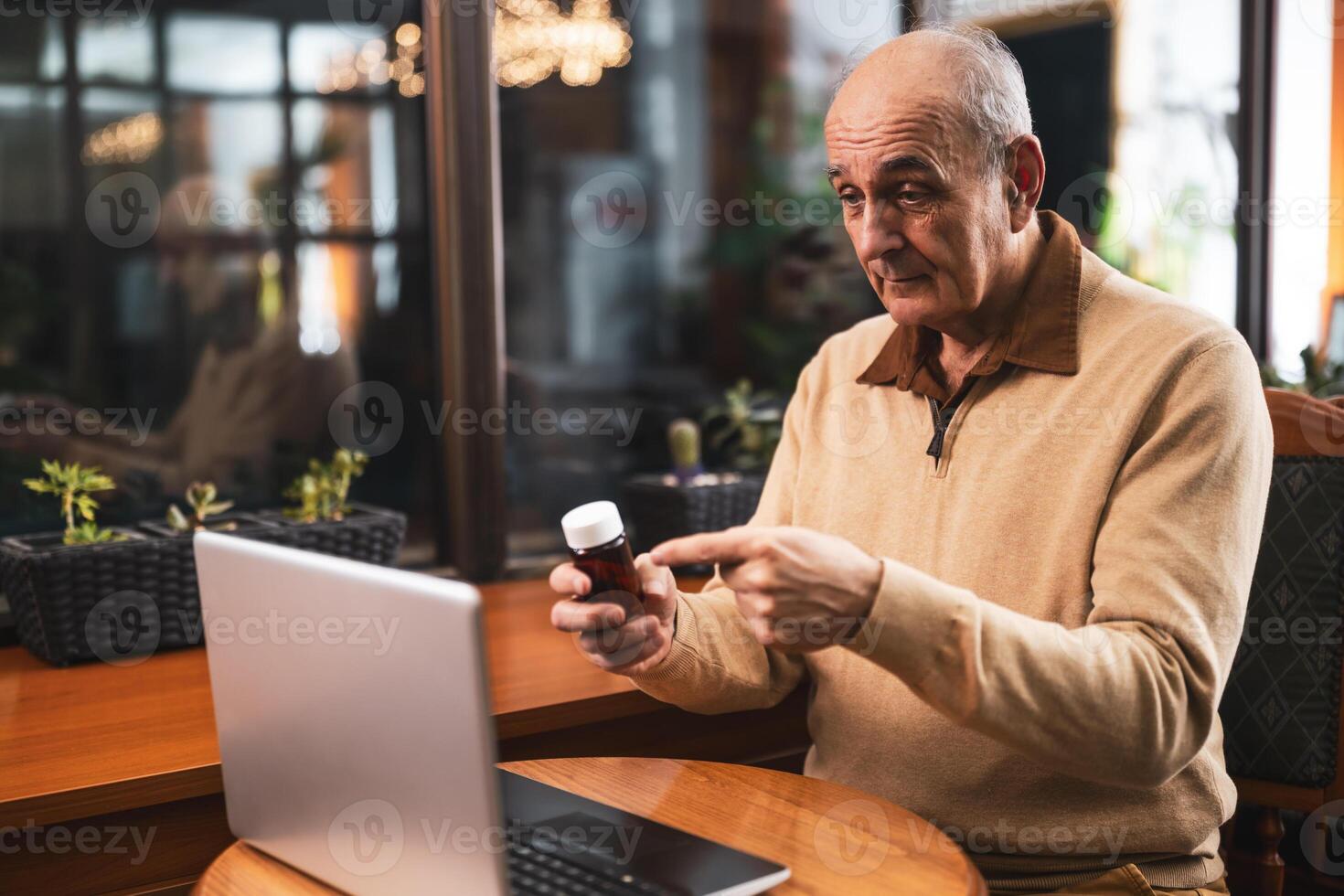 Senior man pointing at bottle of medicine while talking with his doctor on a call via laptop in cafeteria. photo